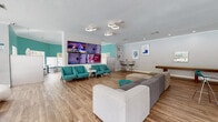 This single-floor community clubhouse is designed with a lively, resort-inspired aesthetic that blends bold color and modern furnishings throughout its open layout. The space is defined by striking teal and cobalt blue accent walls, warm wood-look flooring, and crown molding that adds a polished finish to every room.

The main lounge area serves as the social hub, featuring a large sectional sofa, a wall-mounted multi-screen media display, and a row of retro-style teal accent chairs. Sculptural, organic-form seating pieces in blue and white add an artistic touch and create visual interest throughout the open floor plan. A shuffleboard table and high-top bistro tables with bar stools provide additional options for recreation and casual gathering.

Adjacent to the lounge, a curved wet bar with granite countertops and stylish pendant lighting in deep purple makes a bold design statement, ideal for hosting events. A dedicated dining nook features vibrant teal walls, built-in booth seating, and multiple windows that flood the space with natural light while offering direct views of the outdoor pool area.

Large windows and glass sliding doors frame lush poolside scenery throughout the clubhouse, maintaining a seamless connection between the interior and the outdoor amenities. The overall ambiance is energetic yet comfortable, making this clubhouse a well-equipped and visually dynamic gathering space for residents.