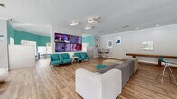 This single-floor community clubhouse is designed with a lively, resort-inspired aesthetic that blends bold color and modern furnishings throughout its open layout. The space is defined by striking teal and cobalt blue accent walls, warm wood-look flooring, and crown molding that adds a polished finish to every room.

The main lounge area serves as the social hub, featuring a large sectional sofa, a wall-mounted multi-screen media display, and a row of retro-style teal accent chairs. Sculptural, organic-form seating pieces in blue and white add an artistic touch and create visual interest throughout the open floor plan. A shuffleboard table and high-top bistro tables with bar stools provide additional options for recreation and casual gathering.

Adjacent to the lounge, a curved wet bar with granite countertops and stylish pendant lighting in deep purple makes a bold design statement, ideal for hosting events. A dedicated dining nook features vibrant teal walls, built-in booth seating, and multiple windows that flood the space with natural light while offering direct views of the outdoor pool area.

Large windows and glass sliding doors frame lush poolside scenery throughout the clubhouse, maintaining a seamless connection between the interior and the outdoor amenities. The overall ambiance is energetic yet comfortable, making this clubhouse a well-equipped and visually dynamic gathering space for residents.