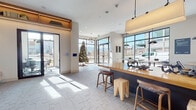 This thoughtfully designed single-floor space blends contemporary aesthetics with functional communal areas. The main lobby and reception zone is immediately welcoming, featuring large floor-to-ceiling windows and glass entry doors that flood the interior with abundant natural light and offer unobstructed views of the surrounding streetscape. Patterned stone tile flooring anchors the open layout, while a substantial dark-base island with a warm butcher block top serves as a central gathering point, complemented by a mix of wood and leather counter stools.

A built-in lounge area along one wall features a upholstered bench with plaid accent pillows beneath illuminated open wood shelving displaying curated books and decorative objects. Adjacent to the lounge, a dedicated nook showcases a textile wall installation framed within an architectural recess, adding an artistic focal point. A compact coffee station with dark cabinetry serves the space nearby.

The conference room offers a distinct atmosphere, distinguished by a textured hexagonal tile accent wall and a wall-mounted display screen. A separate small office or meeting nook with a pendant light and bistro-style table provides additional workspace. Smart home control panels are integrated throughout, reinforcing the space's modern, tech-forward character.