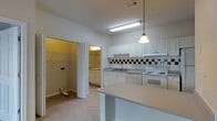 This single-floor space centers around a well-appointed kitchen designed for everyday functionality. White raised-panel cabinetry lines the walls, offering generous storage, while a classic black-and-white tile backsplash adds a subtle decorative detail. The countertops extend into a peninsula-style breakfast bar, providing additional workspace and casual dining potential. A single-basin sink is positioned beneath overhead cabinetry, and white appliances including an electric range with oven and a dishwasher complete the kitchen setup. A pendant light fixture hangs above the peninsula, complementing the flush fluorescent ceiling light to ensure the kitchen is well-illuminated. Adjacent to the kitchen, an open doorway reveals a laundry area with a washer and dryer, adding in-unit convenience. A spacious walk-in closet is also visible nearby, featuring built-in shelving and hanging rods for ample storage. The flooring transitions between tile in the kitchen and carpet in the adjoining areas. Overall, the layout is practical and efficient, making good use of the available space throughout.