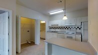 This single-floor space centers around a well-appointed kitchen designed for everyday functionality. White raised-panel cabinetry lines the walls, offering generous storage, while a classic black-and-white tile backsplash adds a subtle decorative detail. The countertops extend into a peninsula-style breakfast bar, providing additional workspace and casual dining potential. A single-basin sink is positioned beneath overhead cabinetry, and white appliances including an electric range with oven and a dishwasher complete the kitchen setup. A pendant light fixture hangs above the peninsula, complementing the flush fluorescent ceiling light to ensure the kitchen is well-illuminated. Adjacent to the kitchen, an open doorway reveals a laundry area with a washer and dryer, adding in-unit convenience. A spacious walk-in closet is also visible nearby, featuring built-in shelving and hanging rods for ample storage. The flooring transitions between tile in the kitchen and carpet in the adjoining areas. Overall, the layout is practical and efficient, making good use of the available space throughout.
