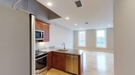 This single-floor residence offers a thoughtfully designed open-concept layout that seamlessly connects the kitchen and living area, creating a bright and functional living environment. The kitchen is outfitted with rich warm-toned cabinetry, granite countertops, and a full suite of stainless steel appliances including a range, built-in microwave, refrigerator, and dishwasher. A peninsula with an undermount sink serves as a natural dividing point between the kitchen and the main living space, offering both additional prep space and a casual gathering spot. A classic subway tile backsplash adds a clean, polished detail to the cooking area. Recessed lighting throughout the ceiling provides consistent, even illumination across the open floor plan. Light hardwood flooring extends seamlessly from the kitchen through the living area, lending warmth and continuity to the space. The living area benefits from generous natural light streaming in through multiple double-hung windows, which frame views of the surrounding neighborhood. Neutral white walls complement the warm wood tones, creating a versatile canvas ready for personalization. The overall ambiance is modern yet inviting, combining practical design with comfortable, everyday livability.