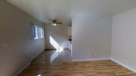This single-floor space offers a clean, functional layout designed for comfortable everyday living. The main living area is filled with natural light streaming through a side window, illuminating the combination of wood-look laminate flooring and tile flooring that transitions between zones. A ceiling fan with a light fixture is centrally positioned overhead, providing both airflow and ambient lighting throughout the room.

The open-concept design connects the living area directly to the kitchen, which is partially separated by a half-wall partition. This architectural detail defines the two spaces while maintaining an open, airy feel. The kitchen area features white cabinetry and appears to include standard appliances, with a dark countertop providing a subtle contrast to the light surroundings.

Throughout the space, the white walls and white baseboards contribute to a bright, neutral palette that creates a sense of openness. The flooring choices — warm wood-look laminate in the living area and stone-style tile in the kitchen transition zone — add visual texture and practicality underfoot. Overall, the space presents a straightforward, well-lit environment with a cohesive design that balances simplicity with functionality.