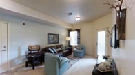 This single-floor living space centers around a thoughtfully arranged living room that balances comfort and functionality. The room features light-toned walls paired with neutral carpeting, creating a warm and inviting atmosphere throughout. A textured ceiling with a central flush-mount light fixture provides consistent overhead illumination, while natural light streams in through two windows dressed with dark patterned curtains, offering flexible light control.

The seating arrangement includes a rich brown leather sofa accented with teal and patterned throw pillows, complemented by a matching teal upholstered armchair that adds a subtle pop of color. A dark wood coffee table anchors the sitting area, which is further defined by a decorative area rug with an intricate pattern. A floor lamp near the window provides additional ambient lighting.

Wall décor includes a framed artwork piece above a dark wood accent table, contributing to the room's curated aesthetic. A door on the right side of the room, featuring a sidelight window, allows additional natural light to filter in and provides access to the exterior, enhancing the overall sense of openness in the space.