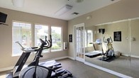 This single-floor fitness center offers a clean, functional environment designed to support an effective workout experience. The space is finished with neutral beige walls and light carpeted flooring, creating a calm and focused atmosphere. Large windows along one wall flood the room with natural light while providing views of the surrounding greenery, enhancing the overall sense of openness. The facility is equipped with a selection of cardio machines, including an elliptical trainer and a treadmill, making it well-suited for a variety of fitness routines. Full-length wall mirrors extend the visual depth of the room and allow users to monitor their form during exercise. A classic French door with a glass panel serves as the entry point, adding a touch of architectural elegance to the otherwise practical space. A wall-mounted informational panel near the entrance provides guidelines for use of the facility. Overhead lighting supplements the natural light, ensuring the space remains bright and well-lit at all times. The overall layout is thoughtfully arranged to maximize usable floor space while maintaining an uncluttered, welcoming environment for residents or guests seeking a convenient on-site fitness option.