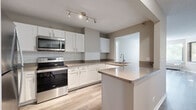 Located at 1101 Iroquois Ave in Naperville, Illinois, this single-floor residence offers a thoughtfully designed layout with a clean, neutral aesthetic throughout. The kitchen is a standout feature, outfitted with crisp white shaker cabinetry, granite-style countertops, a classic subway tile backsplash, and stainless steel appliances including a range and over-the-range microwave. A peninsula with a built-in sink provides additional prep space and defines the kitchen from the adjoining areas. Track lighting illuminates the space, and light wood-look flooring adds warmth underfoot. The living area benefits from a distinctive curved bay window arrangement, flooding the room with natural light and offering pleasant views of surrounding greenery. Light gray wood-look flooring continues through this space, giving it a bright and airy feel. The bedrooms feature plush carpeting, neutral wall tones, and individual windows with views of mature trees outside. Each bedroom includes closet space, and the rooms are well-proportioned for comfortable furnishing. The bathroom is finished with a soaking tub framed in white tile, wood-look flooring, and a separate glass-enclosed shower. A textured ceiling is consistent throughout the home, complementing the understated, move-in-ready character of this Naperville residence.