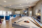 This single-floor community clubhouse offers a thoughtfully designed, multifunctional space that balances recreation, relaxation, and productivity. The open-concept layout is anchored by a central game room featuring a full-size pool table beneath stylish track lighting, with warm wood-look flooring flowing throughout. Soaring ceilings with decorative beam accents and large windows allow abundant natural light to fill the space, offering pleasant views of the surrounding outdoor landscaping.

The lounge area is furnished with bold blue accent chairs, a round coffee table, and a plush area rug, creating an inviting setting for casual gatherings. A wall-mounted flat-screen TV and built-in bench seating add to the entertainment-friendly atmosphere. Adjacent to the lounge, a dining area with a round table and chairs provides a comfortable spot for socializing.

The kitchenette is clean and modern, outfitted with white shaker-style cabinetry, white countertops, stainless steel appliances including a full-size refrigerator and dishwasher, and a coffee station — making it fully functional for community events and everyday use.

A dedicated business center offers two individual workstations with desktop computers, built-in white cabinetry, and tasteful artwork, providing a quiet and productive environment. The overall design palette of warm grey walls, white trim, and rich wood flooring creates a cohesive, contemporary ambiance throughout.