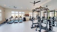 Located at Colonial Gateway Drive in Fort Myers, Florida, this fitness center offers a generously sized, single-floor exercise room designed to support a comprehensive range of workout routines. The space is thoughtfully laid out, with distinct zones dedicated to cardio training, strength work, and functional fitness.

The cardio section is well-appointed with multiple treadmills, elliptical machines, stair climbers, and rowing machines, all positioned along the perimeter to take advantage of the abundant natural light streaming through large banks of windows. These expansive windows line multiple walls, providing wide-open views of the surrounding community and creating a bright, energizing atmosphere throughout.

The strength training area features a full power rack with weight plates, a comprehensive dumbbell rack, and adjustable benches, complemented by full-length mirrors that enhance the sense of openness. A dedicated functional training corner houses TRX suspension equipment, a punching bag, kettlebells, and medicine balls, providing versatility for bodyweight and conditioning workouts.

The interior finishes are clean and modern, with neutral-toned walls, durable speckled flooring, and recessed lighting throughout. Multiple ceiling fans ensure comfortable air circulation. Wall-mounted televisions are positioned for easy viewing from both the cardio and strength zones. Glass double doors provide direct access to the outdoors, further connecting the space to its Florida surroundings.