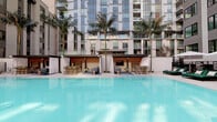 Located at 427 S Hill St in the heart of downtown Los Angeles, this resort-style pool amenity offers a sophisticated outdoor retreat within a modern residential courtyard. The centerpiece is a generously sized swimming pool with crystal-clear turquoise water, complemented by a separate spa tucked into a landscaped alcove with succulent gardens and tiled surrounds. Tall palm trees rise dramatically along the poolside, lending a quintessential California atmosphere to the urban setting. On one side of the pool deck, a row of private cabanas with warm wood-paneled facades and flowing white drapes provide semi-enclosed lounging spaces, each outfitted with seating areas and access to an outdoor TV. On the opposite side, padded chaise lounges in green and gray are arranged beneath large market umbrellas, offering ample space for sunbathing. Glass railings define the perimeter of a secondary lounge terrace, where additional seating is surrounded by native plantings and desert-inspired landscaping. The entire deck is framed by the towering facades of the surrounding residential tower, its floor-to-ceiling glazing and private balconies adding an urban sophistication to the open-air environment. The space seamlessly blends relaxation, style, and function in a vibrant downtown setting.