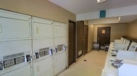 This single-floor shared laundry facility offers a practical and functional space designed to serve the everyday needs of its users. The room is lined with a bank of coin-operated, stacked washer and dryer units positioned along one wall, providing ample laundry capacity. The machines are clearly labeled with operating instructions, making the space straightforward and easy to navigate. The corridor-style layout opens into a wider area toward the back, where two utility room doors provide access to additional building services. The flooring is a light-toned, easy-to-clean surface that runs throughout the space, contributing to a clean and utilitarian aesthetic. Neutral-toned walls keep the environment simple and uncluttered. Toward the far end of the corridor, utility carts are available, offering added convenience for transporting laundry and other items. Overhead lighting ensures the space is well-lit throughout. The overall design prioritizes functionality and accessibility, making this a well-equipped communal laundry room that meets the practical requirements of daily use.