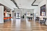 Located at 520 Martin Luther King Jr Blvd in Macon, Georgia, this single-floor community clubhouse is a vibrant, music-themed gathering space that draws inspiration from the city's rich musical heritage. The interior is defined by soaring ceilings, exposed structural beams, and warm hardwood-style flooring that flows continuously throughout the space, creating a cohesive and energetic atmosphere.

At the heart of the lounge is a well-appointed bar area featuring a granite countertop, dark cabinetry, and striking orange upholstered barstools. A dramatic black-and-white mural of a live rock concert spans the back wall, setting a bold, artistic tone. Globe pendant lights hang overhead, adding warmth to the space.

The open lounge area transitions seamlessly into a game room equipped with a full-size billiards table, a wall-mounted flat-screen TV, and a seating nook adorned with large-scale music photography — including an oversized guitar print and classic rock portraiture. Glass-top high tables with modern bar seating are positioned throughout, offering flexible gathering spots.

Large floor-to-ceiling windows and glass doors flood the interior with natural light and provide direct views and access to the outdoor pool area. A glass-enclosed conference or business room is also visible, offering a quieter work setting within the clubhouse. The overall ambiance is modern, bold, and culturally rich.