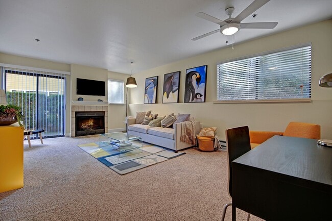 Living room with couch, desk, armchair, large windows, ceiling fan, fireplace, and sliding glass door out to a patio - Foster Greens