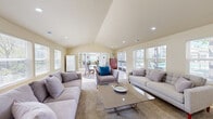 Located at Cobble Creek Circle in Rocklin, California, this community clubhouse offers a welcoming and well-appointed gathering space designed for comfort and socializing. The single-floor layout centers around a spacious living room that immediately impresses with its vaulted, cathedral-style ceiling, which lends a sense of openness and grandeur to the interior.

Natural light is a defining feature throughout, with an abundance of windows fitted with blinds lining multiple walls and allowing views of the surrounding greenery. Recessed lighting complements the natural light, ensuring the space remains bright and inviting at any time of day. The flooring is finished in neutral-toned ceramic tile, providing a clean and durable surface that runs throughout the main area.

The living room is furnished with generous seating arrangements, including plush gray sofas and accent chairs, centered around a glass-top coffee table and area rugs that define distinct lounge zones. A dining area with a round glass table and upholstered chairs provides a casual gathering spot, while a wall-mounted television and media console offer entertainment options.

French doors open directly to an outdoor pool area, seamlessly blending indoor comfort with outdoor leisure. A full-size refrigerator adds convenient amenity functionality. The warm, neutral color palette and thoughtful layout make this clubhouse a practical and pleasant community hub.