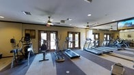This fitness center is designed to support a well-rounded cardio workout in a comfortable, climate-controlled environment. The room features a row of treadmills along one wall, complemented by elliptical trainers and a stationary bike, providing members with a variety of cardio options. A large mirror spans one wall, visually expanding the space and allowing users to monitor their form during exercise.

The room is finished with warm beige walls and patterned carpet flooring in neutral tones, contributing to a clean and welcoming atmosphere. Ceiling fans are installed throughout, offering additional air circulation alongside the HVAC system to keep the space comfortable during workouts.

Natural light filters in through multiple sets of dark-framed glass-panel doors along the back wall, which open to an outdoor area and provide views of the surrounding landscape. Recessed lighting supplements the natural light, ensuring the space remains well-lit at all times. Emergency exit signage is clearly visible, and informational postings are displayed near the entrance doors. Overall, the fitness center offers a practical and inviting environment for daily exercise.