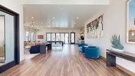 Located on W Lancaster Boulevard in Lancaster, California, this single-story community clubhouse offers a thoughtfully designed and generously proportioned interior. The open-plan entrance and main lounge area are anchored by rich hardwood-style flooring, crisp white walls, and recessed lighting complemented by elegant brass chandelier fixtures that lend a warm, sophisticated glow. Large glass doors along the back wall flood the space with abundant natural light and provide direct views of the outdoor area beyond.

The main lounge is furnished with plush seating, a pool table, and a marble-surround fireplace, creating a versatile environment for both relaxation and socializing. A curated fiber art wall installation adds a striking decorative focal point, while a beverage station provides added convenience for guests.

A private office is accessible via sleek black-framed glass doors, offering a semi-transparent, light-filled enclosure. Inside, a marble-topped round table and teal velvet chairs sit beneath large windows overlooking swaying palm trees. A dramatic Joshua tree mural and slatted wood wall paneling give the room a distinctly Southern California character.

Throughout the space, the design palette of teal, warm neutrals, and natural wood tones creates a cohesive, contemporary ambiance that is both welcoming and refined.