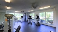 This fitness center is a bright and functional workout facility designed to support a full range of exercise routines. The space is lined with large, multi-pane windows on multiple walls, allowing abundant natural light to flood the interior and offering pleasant views of the surrounding greenery. The open floor plan provides ample room for movement between equipment, which includes several elliptical cardio machines, a cable pulley strength training system, a dumbbell rack, and a workout bench. Dark rubberized flooring covers the entire area, offering durability, traction, and comfort underfoot. The ceiling features a textured finish and is equipped with two ceiling fans that promote airflow throughout the room, complemented by fluorescent strip lighting for consistent illumination during any hour. The white walls contribute to the clean, airy atmosphere of the space. The combination of natural light, well-maintained equipment, and thoughtful layout creates a welcoming and practical environment suited for both cardiovascular and strength-based training.