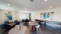 This well-appointed game and lounge space is designed for social recreation, offering a diverse range of entertainment options within a contemporary, open layout. The flooring throughout features a striking dark herringbone-patterned tile that anchors the room's modern aesthetic and provides a cohesive visual flow from one zone to the next.

The gaming area is a standout highlight, housing a full-size pool table illuminated by bold cylindrical pendant lights, a classic shuffleboard table displayed beneath a series of colorful abstract wall panels, and a dedicated poker table set beneath an eye-catching geometric chandelier. Each gaming zone is thoughtfully defined while remaining open to the broader space.

A comfortable lounge area with navy blue accent chairs and small side tables offers a relaxed setting for conversation, while multiple round dining-height tables with modern chairs provide additional flexible seating. A wall-mounted flat-screen television and built-in cabinetry add functional amenities to the pool table area.

Natural light filters in through multiple windows with clean white blinds, brightening the neutral white walls. French doors open to an outdoor patio area, extending the social atmosphere beyond the interior. The overall ambiance is modern, inviting, and well-suited for communal gatherings.
