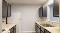 This single-floor residential unit offers a practical and comfortable layout designed for everyday living. The galley-style kitchen is well-appointed with dark charcoal cabinetry, white countertops, a double-basin stainless steel sink, an electric coil range, a refrigerator, and an overhead microwave. A pass-through window connects the kitchen to an adjacent area, enhancing the sense of openness. Wood-look luxury vinyl plank flooring runs throughout the kitchen and entry areas, providing a clean, modern aesthetic.

The entry hallway leads directly into the main living spaces, where the flooring transitions to hardwood-style planks in a cool grey tone. The living area benefits from natural light streaming through a window fitted with blinds, and includes a wall-mounted air conditioning unit and baseboard heating. A carpeted bedroom features a ceiling fan with light fixture, a window with blinds, and baseboard heating, creating a warm and cozy retreat. An additional carpeted room offers flexible use and connects directly to the kitchen.

The full bathroom is finished with a tub-shower combination, a white vanity with sink, a toilet, and a large mirror, all set on wood-look flooring. A fire extinguisher is visibly mounted in the kitchen area for safety. The unit presents a clean, functional space suitable for comfortable residential living.