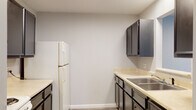 This single-floor residential unit offers a practical and comfortable layout designed for everyday living. The galley-style kitchen is well-appointed with dark charcoal cabinetry, white countertops, a double-basin stainless steel sink, an electric coil range, a refrigerator, and an overhead microwave. A pass-through window connects the kitchen to an adjacent area, enhancing the sense of openness. Wood-look luxury vinyl plank flooring runs throughout the kitchen and entry areas, providing a clean, modern aesthetic.

The entry hallway leads directly into the main living spaces, where the flooring transitions to hardwood-style planks in a cool grey tone. The living area benefits from natural light streaming through a window fitted with blinds, and includes a wall-mounted air conditioning unit and baseboard heating. A carpeted bedroom features a ceiling fan with light fixture, a window with blinds, and baseboard heating, creating a warm and cozy retreat. An additional carpeted room offers flexible use and connects directly to the kitchen.

The full bathroom is finished with a tub-shower combination, a white vanity with sink, a toilet, and a large mirror, all set on wood-look flooring. A fire extinguisher is visibly mounted in the kitchen area for safety. The unit presents a clean, functional space suitable for comfortable residential living.