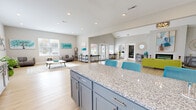 This single-floor community clubhouse offers a generously sized, open-concept layout designed for comfort and social interaction. The space is anchored by a dramatic vaulted ceiling in the main lounge area, which draws the eye upward and creates an airy, expansive atmosphere. Light hardwood flooring runs throughout, complementing the neutral gray and white color palette accented with vibrant teal and chartreuse furnishings.

The main lounge features a sleek linear fireplace, a stylish pendant chandelier, and large windows that flood the space with natural light. Comfortable seating arrangements, including accent chairs and a bold lime-green sofa, are positioned around patterned area rugs, creating inviting conversation areas. Wall art in aqua and abstract tones adds a contemporary decorative touch.

Adjacent to the lounge, a well-appointed kitchen area includes gray cabinetry with granite countertops, a central island with bar seating, a stainless steel refrigerator, and a sink — ideal for community gatherings and events. A dedicated coffee bar station further enhances the hospitality of the space.

A quieter seating nook with large windows offers a more relaxed retreat, and a separate fitness center is accessible via interior windows and a connecting door, rounding out this versatile and welcoming community space.