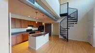 Located at 425 Macdougall St in Columbia, South Carolina, this two-story loft-style space showcases an industrial-modern aesthetic defined by soaring double-height ceilings and exposed metallic ductwork running prominently across the open main level. The centerpiece of the lower floor is a striking black wrought-iron spiral staircase that connects both levels, serving as both a functional element and a bold architectural statement.

The open-concept kitchen features warm-toned wood cabinetry with dark countertops, black appliances including a range and refrigerator, and a generously sized island with a built-in sink and pendant lighting overhead. Track lighting complements the pendants, ensuring ample illumination throughout. Light hardwood flooring extends across the main living areas, creating a bright, airy feel. Large windows and a sliding glass door bring in natural light and offer views of the surrounding greenery.

The upper level, accessed via the spiral staircase, offers open carpeted space with an open-railing overlook, exposed ductwork, and windows admitting natural light. A bedroom on this level features plush carpet flooring and a spacious closet. The bathroom is finished with dark tile flooring, wood cabinetry, and a well-lit vanity. A sliding barn door adds a contemporary industrial touch to the overall design.