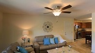 This single-floor residence offers a comfortable and inviting living environment designed with everyday functionality in mind. The living area serves as the heart of the home, featuring warm, neutral-toned walls that create a relaxed and welcoming atmosphere. A plush leather sofa accented with vibrant teal throw pillows provides ample seating, while a light wood coffee table anchors the space. A classic five-blade ceiling fan with an integrated light fixture hangs overhead, ensuring year-round comfort and steady ambient illumination throughout the room. Decorative touches, including an ornate metal wall accent and a softly lit table lamp, add character and warmth to the space. Light-toned flooring flows throughout, contributing to an open and airy feel. The layout is practical and well-proportioned, offering a natural flow between areas. Interior doors with clean, white trim provide access to additional rooms, reflecting a straightforward and tidy architectural style. Overall, this home presents a cozy, move-in ready environment that balances comfort with understated charm, making it an ideal space for everyday living and relaxation.