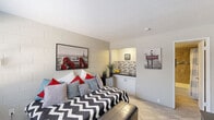 This compact single-floor unit offers a thoughtfully organized layout that maximizes every square foot across one bedroom and one full bathroom. The main living area features light-toned ceramic tile flooring and exposed concrete block walls painted in a clean white finish, lending the space a sturdy, no-frills character. A wall-mounted flat-screen television and a low-profile dresser unit are positioned along one wall, while a window with blinds allows natural light to filter in and accommodates a wall-mounted AC unit for climate control. The sleeping area flows seamlessly into a recessed kitchenette alcove, which is equipped with white cabinetry, a dark countertop, and a mosaic tile backsplash — providing essential functionality within a minimal footprint. Paris-themed artwork displayed throughout the space adds a cohesive and stylish decorative element. The bathroom features warm-toned large-format floor and wall tiles, a soaking tub with a shower combo, a mosaic accent tile border, and a built-in niche for storage. The overall ambiance is clean, practical, and purposefully styled for efficient single-occupant living.