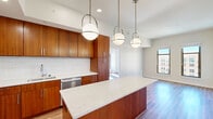 This thoughtfully designed 2-bedroom, 2-bathroom residence on a single floor offers a cohesive blend of modern aesthetics and functional living. The open-concept kitchen is a standout feature, showcasing rich warm-toned wood cabinetry, a white subway tile backsplash, quartz countertops with subtle veining, and a large center island ideal for dining or prep work. A full suite of stainless steel appliances — including a gas range, microwave, refrigerator, and dishwasher — completes the culinary space. Pendant globe lights with brushed nickel accents illuminate the kitchen and flow naturally into the adjacent living area, which benefits from abundant natural light through large windows offering urban skyline views. Warm hardwood-style flooring runs throughout the living spaces and bedrooms, adding continuity and warmth. The primary bedroom is generously sized and features dual windows with city views and access to a dedicated closet. The second bedroom also enjoys natural light and outdoor views. Both bathrooms are finished in a clean, contemporary style — one features a glass-enclosed walk-in shower with white tile, while the other includes a full tub/shower combo, warm wood vanity, and large-format tile flooring. The overall ambiance is bright, clean, and modern throughout.