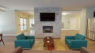Located in Castle Shannon, PA, this single-floor community space offers a thoughtfully designed layout ideal for gathering and relaxation. The centerpiece of the main lounge is a striking floor-to-ceiling stacked stone fireplace wall, complete with a mounted flat-screen TV, flanked by teal mid-century modern sofas arranged over a vibrant area rug — creating a warm and inviting focal point. Durable wood-look flooring runs throughout, unifying the open layout. Multiple seating vignettes line the perimeter, each positioned near large windows with horizontal blinds that invite abundant natural light and views of the surrounding area. The soft sage and warm beige wall tones contribute to a calm, welcoming atmosphere. A dedicated dining area features round wood tables with coordinating chairs, offering comfortable spaces for casual meals or group conversations. The kitchenette is outfitted with dark espresso cabinetry, white subway tile backsplash, light quartz countertops, a stainless steel sink, and a full-size stainless steel refrigerator — providing practical amenities for residents and guests. A built-in mailbox wall and accessible entry doors add functional convenience. The overall design balances modern aesthetics with community-focused comfort.