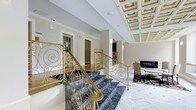 This sophisticated space makes a striking first impression with its grand marble staircase, finished in deep grey veining and flanked by elaborately scrolled gold wrought-iron railings that serve as a defining architectural centerpiece. The craftsmanship of the metalwork is exceptionally detailed, featuring flowing scroll patterns that complement the overall classical design language of the space. Overhead, a striking coffered ceiling with intricate geometric paneling and decorative molding adds depth and a sense of grandeur, drawing the eye upward and reinforcing the formal character of the interior. The flooring transitions from the bold marble stairs to light-toned tile, contributing to a bright and airy atmosphere. Neutral, warm-toned walls provide a refined backdrop that allows the architectural elements to stand out. Adjacent to the staircase, a seating area with upholstered chairs is anchored by a patterned area rug and positioned near a fireplace, offering an inviting retreat. Additional doorways visible throughout suggest further rooms beyond, maintaining a sense of openness and connectivity. The overall ambiance is one of timeless elegance and classical formality.
