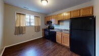This single-floor residence offers a straightforward and functional layout designed for comfortable everyday living. The floor plan includes multiple bedrooms, a kitchen, and a full bathroom, all connected by a practical and efficient arrangement of rooms.

The bedrooms are finished with neutral-toned carpet flooring and smooth, light-colored walls, creating a clean and understated atmosphere. Each room benefits from natural light through windows, with some featuring multi-pane windows that allow views of surrounding greenery. Built-in closet storage is present in at least one bedroom, adding to the unit's practical appeal.

The kitchen is well-equipped, featuring warm wood-toned cabinetry, dark granite-style countertops, and a full suite of black appliances including an electric range, overhead ventilation hood, and refrigerator. A built-in sink and a window with outdoor views round out this functional cooking space. Dark hardwood-style flooring ties the kitchen together with a cohesive, durable finish.

The bathroom is neatly appointed with a tub and shower combination enclosed by sliding frosted glass doors with chrome framing. Wood-style flooring continues here, complementing the clean white fixtures including the toilet and tub surround. The overall ambiance of the space is practical and move-in ready, with consistent neutral tones and durable finishes throughout.