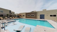 This outdoor amenity space centers around a generously sized rectangular swimming pool with clear, blue water, designed for leisure rather than competitive swimming, as indicated by the no-diving designation. The pool deck is finished in a clean, light-toned concrete surface and is equipped with multiple cushioned lounge chairs and chaise lounges, providing ample space for sunbathing and relaxation. A red patio umbrella adds a pop of color to the seating area, offering shaded respite on sunny days. The surrounding area is enclosed by a black metal fence, ensuring safety and privacy for users. A well-maintained clubhouse or amenity building anchors the space, featuring a combination of brown wood-panel and tan siding exterior, multiple windows, and secure double doors — suggesting restroom or changing facilities. Wall-mounted lighting fixtures on the building provide illumination for evening use. The backdrop reveals a residential community with additional buildings visible beyond the fence line. The overcast sky lends the space a calm, neutral ambiance, while the overall layout reflects a thoughtfully designed community amenity built for comfort, safety, and social enjoyment.