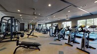 This contemporary fitness center is thoughtfully designed to accommodate a wide range of workout needs across a single, expansive floor. The main gym area features a herringbone-patterned carpet in neutral tones with teal accents, providing a clean and modern aesthetic underfoot. The space is outfitted with an extensive selection of strength machines, cable equipment, free weights including kettlebells, and a full lineup of cardio equipment — including treadmills, stationary bikes, ellipticals, and stair climbers. Ceiling fans and recessed lighting ensure a comfortable, well-lit environment, while large windows along the perimeter flood the space with natural light and offer views of lush, tree-lined outdoor surroundings. A dedicated group exercise and yoga studio is finished with rich wood-look flooring, gold accent lighting, a wall-mounted TV, a ballet barre, exercise balls, and spin bikes — ideal for classes or individual training. An adjoining room with plush carpet, floor-to-ceiling windows, and a wall-mounted TV serves as a children's activity area. Additional amenities include water fountains, open shelving for equipment storage, restrooms, and glass-panel dividers that maintain visual connectivity throughout the facility. The overall ambiance is modern, airy, and inviting.
