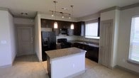 This single-floor space centers around a well-appointed kitchen designed for both functionality and visual appeal. The kitchen features dark espresso cabinetry that extends to the upper and lower walls, providing ample storage throughout. Granite countertops with a speckled beige-and-gray pattern run along the perimeter and top the central island, which serves as a versatile workspace or casual dining area with a clean white base.

Black appliances, including a refrigerator with an ice and water dispenser, a range, a built-in microwave, and a dishwasher, complement the dark cabinetry for a cohesive aesthetic. A stainless steel sink is positioned beneath a window fitted with white blinds, allowing natural light to filter into the space.

Overhead, a combination of track lighting and two pendant lights with frosted glass shades provides layered illumination across the kitchen. The flooring transitions from tile in the kitchen area to carpet in the adjoining space, subtly delineating the zones. A door on the far right leads to an adjacent room, adding to the practical layout of this well-designed interior.