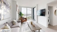 This single-floor residence is designed with a contemporary aesthetic, characterized by light wood-style flooring, crisp white walls, and expansive floor-to-ceiling windows that frame striking panoramic views of the city skyline and nearby waterway. The open-plan living area is bright and airy, with large glass panels allowing abundant natural light to fill the space. The kitchen is outfitted with dark wood cabinetry, white countertops, and a full suite of stainless steel appliances, including a refrigerator, range, and built-in microwave, all set against light flooring for a clean, modern contrast. The primary bedroom benefits from wraparound floor-to-ceiling windows on multiple walls, providing sweeping elevated views of the surrounding cityscape and water. It is complemented by a dedicated walk-in closet featuring custom built-in shelving and drawers for generous storage. The bathrooms are finished with large-format stone-look tiles and frameless glass-enclosed walk-in showers equipped with rainfall showerheads and integrated lighting. The overall ambiance throughout is polished and contemporary, with an emphasis on natural light and urban views.