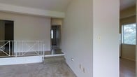 This single-floor space presents a clean, minimalist aesthetic with neutral wall tones and a notable split-level layout that adds architectural interest. A raised platform section, accessible via a low step, divides the interior into distinct zones, creating a sense of depth and dimension despite the compact footprint. A white metal railing borders the raised area, providing both a functional and decorative element that draws the eye across the space. The flooring transitions between what appears to be tile or stone on the raised platform and a softer carpeted surface on the lower level, offering textural contrast. A hallway visible in the background leads to additional areas, with a doorway at its end hinting at natural light from a window beyond. The walls are smooth and painted in soft, neutral tones throughout, contributing to a calm and understated ambiance. Electrical outlets and light switch plates are positioned along the walls, and the overall condition of the space appears ready for furnishing or renovation. The layout provides a functional flow between the different levels and connected rooms.