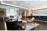 Located in the heart of Boise, ID, this beautifully appointed lobby offers a spacious and refined welcome experience across a single, generously sized floor. The design blends contemporary sophistication with warm, natural materials to create an inviting yet polished atmosphere.

At the center of the space is a striking navy-blue reception bar topped with a light marble surface, lined with upholstered bar-height seating and set atop a geometric hexagonal tile inlay — a standout design feature that anchors the room. Behind the bar, an artistic wall installation of scattered, colorful sculptural elements adds a creative focal point.

Rich, dark hardwood flooring runs throughout, complemented by warm walnut wood wall paneling that lends a sense of depth and texture. Recessed lighting and subtle cove illumination along the ceiling provide a layered, ambient glow throughout the hallway and lounge areas.

A comfortable lounge zone features mid-century modern seating arranged around a fireplace, offering an intimate gathering area. The entrance is defined by floor-to-ceiling glass curtain walls and a revolving door that floods the space with natural light and provides clear views of the surrounding Boise streetscape. Pendant lighting near the entry adds a warm, decorative touch, completing the lobby's refined and welcoming character.