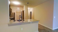 This single-floor residence showcases a practical and open-concept design that seamlessly connects the kitchen and living areas. The kitchen is anchored by a peninsula counter finished in granite, offering ample prep space and featuring an integrated stainless steel sink with a modern gooseneck faucet. The peninsula also provides convenient electrical outlets, adding functional versatility to the space. A stainless steel refrigerator is visible along the kitchen wall, complementing the clean, contemporary aesthetic.

Wood-style flooring extends throughout the main living area, providing a warm and durable surface underfoot. The walls are finished in a neutral warm beige tone, contributing to an inviting and cohesive atmosphere. Flush-mount ceiling light fixtures provide consistent overhead illumination throughout the space.

The open layout offers clear sightlines from the kitchen into the living area and hallway beyond, where multiple doorways lead to additional rooms. The overall design emphasizes functional flow between spaces, with clean lines and neutral finishes that create a versatile environment suitable for a variety of furnishing styles.