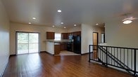 This two-story residence is designed with an open-concept layout on the main level, seamlessly connecting the living area and kitchen. The kitchen is well-appointed with dark wood cabinetry, dark granite-style countertops, and black appliances including a refrigerator, stove, and built-in microwave. A raised breakfast bar provides a casual dining option and acts as a natural divider between the kitchen and the open living space. Recessed lighting throughout the ceiling provides consistent, even illumination across the main level. The hardwood floors extend across the living and kitchen areas, adding warmth and continuity to the space, while tile flooring defines the kitchen work area. A sliding glass door on one side of the main level opens to a balcony, inviting natural light and outdoor views into the interior. A staircase with a metal railing leads to the upper level, accompanied by a hallway that connects to additional rooms. The neutral wall tones throughout complement the darker wood finishes, creating a balanced and functional living environment.
