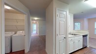 This single-floor residence offers a thoughtfully designed layout that balances functionality and comfort throughout. The open-concept kitchen flows seamlessly into the living area, featuring white cabinetry, stainless steel appliances including a gas range and refrigerator, and a peninsula counter that provides both additional workspace and a natural division between the kitchen and the carpeted living space. Warm-toned carpet extends throughout the living and bedroom areas, creating a cohesive and inviting atmosphere.

A central hallway connects the main living spaces to the private areas of the home, including the bedrooms and bathrooms. The bedroom features plush carpeting, a sliding closet, and corner windows that allow natural light to fill the room while offering views of the surrounding greenery. Two full bathrooms are finished consistently with white vanity cabinetry, light countertops, tile flooring, full tub-shower combinations, and large mirrors with overhead lighting.

A dedicated in-unit laundry area houses a full-size washer and dryer set, complete with overhead wire shelving for additional storage. The neutral color palette of warm beige and white tones throughout the space contributes to a clean, bright, and move-in-ready environment well-suited for comfortable everyday living.