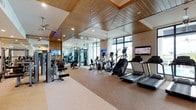 This modern fitness center offers a spacious, single-floor layout designed to support a full range of workout routines. The space is immediately distinguished by its striking wood-paneled ceiling accents, recessed lighting, and ceiling fans that lend a warm, resort-style ambiance while maintaining a comfortable environment for exercise.

The cardio zone is lined with treadmills, elliptical trainers, stair climbers, and stationary bikes, many equipped with individual TV screens for entertainment during workouts. Floor-to-ceiling windows along one wall flood the space with abundant natural light and provide views of an outdoor pool and lush landscaping, creating an energizing atmosphere.

The strength training area is generously outfitted with a wide array of selectorized cable machines, weight benches, and resistance equipment from brands including Matrix. A dedicated dumbbell rack with a full range of free weights occupies a central station, complemented by wall-to-wall mirrors that enhance the sense of openness and allow users to monitor their form.

Neutral-toned carpet flooring defines the main exercise areas, providing comfort underfoot. Multiple flat-screen TVs are mounted throughout the space, ensuring entertainment is accessible from virtually any station. The overall design balances functionality with aesthetic appeal, delivering a polished, professional fitness environment.