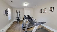This fitness center offers a clean, functional workout environment designed to support a variety of exercise routines. The space is finished with warm hardwood-style flooring throughout, providing a durable and attractive surface suited for both cardio and strength training activities. Neutral-toned walls and flush-mounted ceiling lighting create a bright, welcoming atmosphere conducive to focused exercise sessions.

The cardio area is anchored by two True-brand treadmills, each equipped with digital displays for tracking workout metrics. Complementing the cardio equipment is a multi-function cable machine with an adjustable bench, allowing users to perform a wide range of strength and resistance exercises. A stability ball is also present, adding options for core and flexibility training.

Natural light filters into the room through windows fitted with horizontal blinds, offering ventilation and a connection to the outdoors while maintaining privacy. Motivational framed prints are displayed along the walls, adding a purposeful and energizing aesthetic to the space. A paper towel dispenser is conveniently mounted on the wall, supporting hygiene and equipment maintenance. Access to the fitness center is provided through a glass-paneled entry door as well as an interior door, ensuring easy and convenient entry and exit for all users.