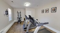 This fitness center offers a clean, functional workout environment designed to support a variety of exercise routines. The space is finished with warm hardwood-style flooring throughout, providing a durable and attractive surface suited for both cardio and strength training activities. Neutral-toned walls and flush-mounted ceiling lighting create a bright, welcoming atmosphere conducive to focused exercise sessions.

The cardio area is anchored by two True-brand treadmills, each equipped with digital displays for tracking workout metrics. Complementing the cardio equipment is a multi-function cable machine with an adjustable bench, allowing users to perform a wide range of strength and resistance exercises. A stability ball is also present, adding options for core and flexibility training.

Natural light filters into the room through windows fitted with horizontal blinds, offering ventilation and a connection to the outdoors while maintaining privacy. Motivational framed prints are displayed along the walls, adding a purposeful and energizing aesthetic to the space. A paper towel dispenser is conveniently mounted on the wall, supporting hygiene and equipment maintenance. Access to the fitness center is provided through a glass-paneled entry door as well as an interior door, ensuring easy and convenient entry and exit for all users.