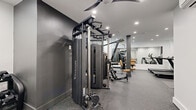 Located at 100 Tuers in Jersey City, NJ, this fitness center offers a thoughtfully designed workout space that maximizes its footprint to deliver a full-featured gym experience. The interior features a clean, modern aesthetic with a neutral color palette of white and dark gray walls, speckled rubber flooring for durability and safety, and recessed ceiling lighting that keeps the space bright and functional throughout.

The gym is outfitted with a diverse selection of equipment to support a complete workout regimen. Cardio options include multiple treadmills and an elliptical trainer, while strength training is well-represented with a Vision functional trainer cable machine, a full rack of Hoist selectorized machines, a dumbbell rack with a wide range of free weights, and a flat utility bench. Large wall-mounted mirrors enhance the sense of space and allow users to monitor their form during exercise.

Structural columns are integrated neatly into the layout without disrupting traffic flow, and a ceiling fan aids in ventilation and comfort. High, narrow windows along one wall allow natural light to filter in while maintaining privacy. A dedicated area with a wall-mounted split HVAC unit ensures the space remains climate-controlled year-round. The overall design is efficient, modern, and purpose-built to provide residents with a convenient and fully equipped place to train.