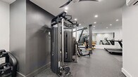 Located at 100 Tuers in Jersey City, NJ, this fitness center offers a thoughtfully designed workout space that maximizes its footprint to deliver a full-featured gym experience. The interior features a clean, modern aesthetic with a neutral color palette of white and dark gray walls, speckled rubber flooring for durability and safety, and recessed ceiling lighting that keeps the space bright and functional throughout.

The gym is outfitted with a diverse selection of equipment to support a complete workout regimen. Cardio options include multiple treadmills and an elliptical trainer, while strength training is well-represented with a Vision functional trainer cable machine, a full rack of Hoist selectorized machines, a dumbbell rack with a wide range of free weights, and a flat utility bench. Large wall-mounted mirrors enhance the sense of space and allow users to monitor their form during exercise.

Structural columns are integrated neatly into the layout without disrupting traffic flow, and a ceiling fan aids in ventilation and comfort. High, narrow windows along one wall allow natural light to filter in while maintaining privacy. A dedicated area with a wall-mounted split HVAC unit ensures the space remains climate-controlled year-round. The overall design is efficient, modern, and purpose-built to provide residents with a convenient and fully equipped place to train.