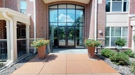 This single-story commercial building makes a strong first impression with its well-crafted brick exterior and a prominent, floor-to-ceiling glass curtain wall entrance. The grand entryway is framed by a sweeping arched transom and fitted with sleek dark-framed glass panels, flooding the entry vestibule with natural light and offering a clear visual connection between the interior and the outdoors. Double glass doors provide welcoming access, flanked on either side by large terra cotta-toned planters filled with vibrant orange flowering plants, adding a warm and inviting touch to the approach. The exterior facade combines classic red brick masonry with refined architectural accents, including a subtly bowed entry overhang that lends a sense of formality and permanence to the building's presence. A wide, paved entry plaza provides ample arrival space, finished in earthy tones that complement the overall color palette of the structure. The building number is prominently displayed near the entrance, reinforcing its professional character. The interior, while minimally lit in the available imagery, suggests a series of enclosed rooms or corridors behind the luminous facade. Overall, the building presents a polished, professional aesthetic well-suited for commercial or institutional use.