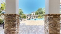 Located at 114 Estuary Trail in Alpharetta, Georgia, this single-level outdoor amenity space serves as a resort-style pool complex designed for leisure and relaxation. The centerpiece is a large, rectangular swimming pool with clear blue water, marked at a maximum depth of 5 feet with a no-diving designation, ensuring a safe and inviting environment for residents.

Surrounding the pool is an expansive herringbone-patterned brick patio that provides ample space for outdoor activity and socializing. Rows of modern chaise lounge chairs are arranged along the pool deck, offering plenty of sunbathing opportunities. Additional seating areas and tables are distributed throughout the space for comfort and convenience.

A covered poolside cabana or pavilion structure anchors one side of the space, featuring stone veneer column accents and a pitched roof that provides shaded seating underneath. On the opposite end, a wood pergola offers another shaded retreat framed by mature trees and tropical-style plantings, including palms and lush shrubs.

The entire area is enclosed by decorative iron fencing and enveloped by a backdrop of towering pine trees and dense greenery, creating a private, natural atmosphere. The surrounding residential buildings are visible in the background, reinforcing the community-oriented nature of this well-appointed outdoor amenity space.