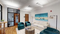 The Resident Lounge is a thoughtfully designed communal space that offers a comfortable and inviting atmosphere for relaxation and socializing. The room features a clean, modern aesthetic with white walls and a drop-tile ceiling equipped with bright linear lighting fixtures that ensure a well-lit environment throughout the day.

The seating area is tastefully furnished with a plush white sofa and a pair of rich teal velvet accent chairs, arranged around a rustic circular wooden coffee table. A geometric-patterned area rug in white and gold anchors the seating arrangement, adding a touch of sophistication to the space. A statement floor lamp provides additional ambient lighting.

Two vibrant teal and cream abstract canvas paintings adorn the wall, serving as a bold focal point that ties together the room's color palette. Warm hardwood-style flooring extends throughout, adding warmth and visual continuity to the space.

Access to adjoining areas is provided through wood-toned interior doors, while a glass-paneled entry door with sidelights allows natural visibility into the corridor. The lounge is designed to serve as a functional yet stylish gathering place, balancing practicality with comfort to create an appealing environment for residents.