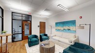 The Resident Lounge is a thoughtfully designed communal space that offers a comfortable and inviting atmosphere for relaxation and socializing. The room features a clean, modern aesthetic with white walls and a drop-tile ceiling equipped with bright linear lighting fixtures that ensure a well-lit environment throughout the day.

The seating area is tastefully furnished with a plush white sofa and a pair of rich teal velvet accent chairs, arranged around a rustic circular wooden coffee table. A geometric-patterned area rug in white and gold anchors the seating arrangement, adding a touch of sophistication to the space. A statement floor lamp provides additional ambient lighting.

Two vibrant teal and cream abstract canvas paintings adorn the wall, serving as a bold focal point that ties together the room's color palette. Warm hardwood-style flooring extends throughout, adding warmth and visual continuity to the space.

Access to adjoining areas is provided through wood-toned interior doors, while a glass-paneled entry door with sidelights allows natural visibility into the corridor. The lounge is designed to serve as a functional yet stylish gathering place, balancing practicality with comfort to create an appealing environment for residents.