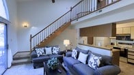 This two-story space makes a strong first impression with its soaring ceilings and an elegant wooden staircase that serves as a central architectural focal point. The staircase features warm-toned wood balusters and a classic railing design, rising to an open upper-level overlook that adds depth and visual interest to the main living area below.

The main floor living area is generously proportioned, accommodating a pair of coordinated gray sofas adorned with decorative patterned and solid pillows, centered around a rectangular coffee table. The layout encourages comfortable conversation and relaxed living. Neutral-toned carpeting grounds the space, while white walls reflect light and contribute to the open, airy atmosphere created by the high ceilings.

Adjacent to the living area, a pass-through opening reveals a glimpse of the kitchen, which features warm wood cabinetry and a clean, functional layout. The upper level, visible from below through the wood railing system, adds a sense of connectivity between floors while maintaining a feeling of spaciousness throughout. Overall, the space balances classic design elements with a comfortable, livable aesthetic.